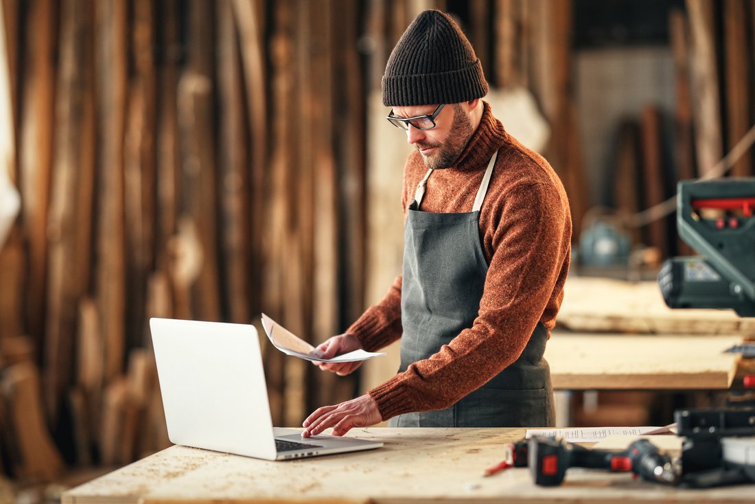 Handwerksunternehmer angespannt zwischen Laptop und Papierkram in der Werkstatt
